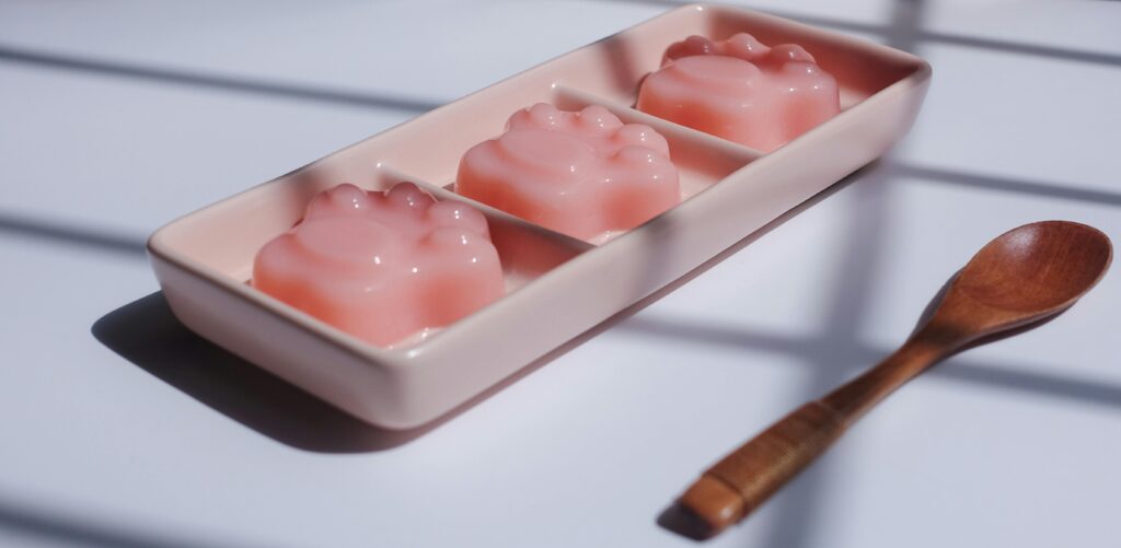 Three pink jelly desserts in a tray, accompanied by a wooden spoon on a sunlit table.